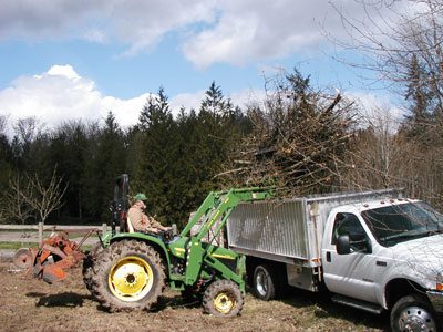 A tractor providing posthole digging services by pulling a truck with a tree in the back.